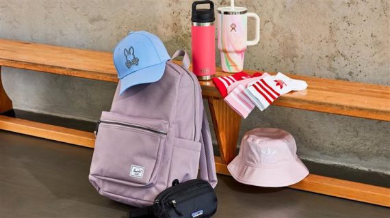 a wooden bench with a light purple/greyish Herschel backpack in front, a blue Psycho Bunny baseball cat on top of the backpack, a light pink adidas bucket hat below the bench, to the right of the backpack; a pink YETI tumbler on top of the bench next to a light pink Stanley straw tumbler and 3 different adidas crew socks