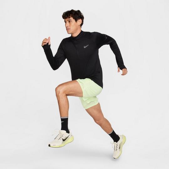 male model in a running action position wearing a Nike black half-zip long sleeve top with a Nike Swoosh logo at the left chest, pale yellow/green running shorts with black crew-length socks and white/yellow Nike running shoes; gym class essentials
